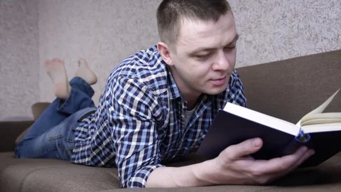 Funny man reading book while lying on couch at home. Knowledge and learning Stock Footage 129663355