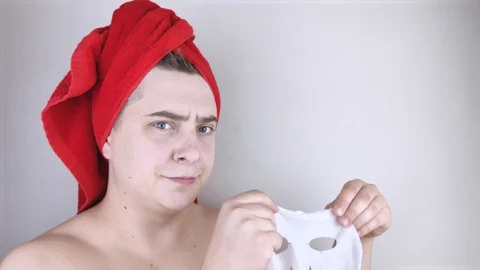 Funny man with a red towel puts a moisturizing fabric mask on his face. The conc Video stock 128362782