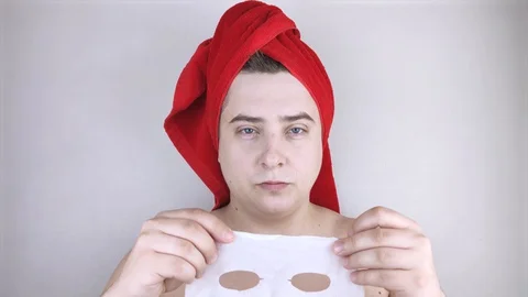 Funny man with a red towel puts a moisturizing fabric mask on his face. The conc Video stock 128362807