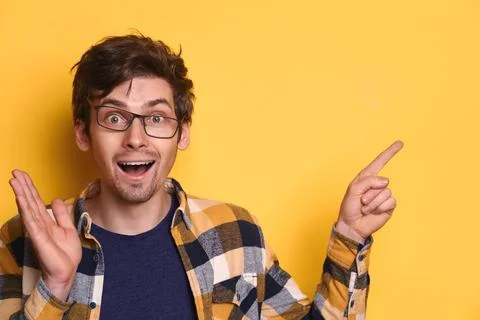 Funny nerd guy looks amazed and shows something on yellow studio wall Foto stock