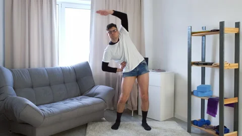 Funny nerd man in glasses and shorts doing complex of fitness exercises at home. Video stock 153356596