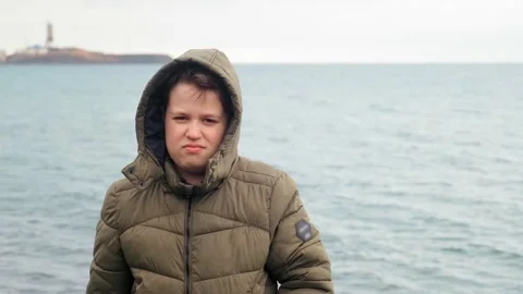 Funny schoolboy in brown jacket poses for camera against sea 库存影片 134268059