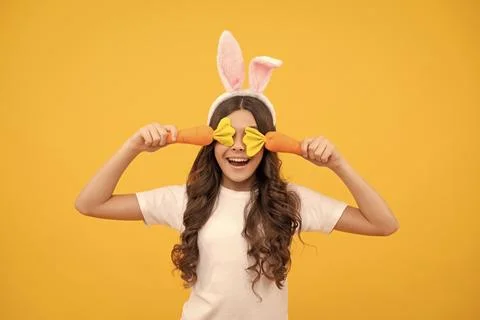 Funny teen girl in bunny ears hold carrot on yellow background, easter Stock Photos