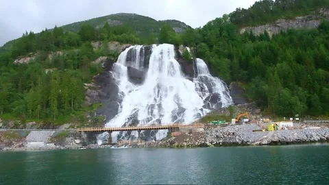 The Furebergfossen Waterfall runs down mountain with pines and bridge building Stock Footage 168371619