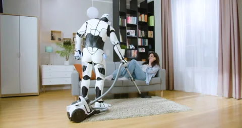Futuristic cyborg using vacuum cleaner while woman in casual wear lying on comfy Stock Footage 244140094