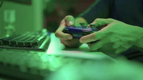 Gamer gripping red controller in dark hoodie, lowlit room with neon green PC and Stock Footage 347874762