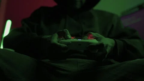 Gamer gripping red controller in dark hoodie, lowlit room with neon green PC and Stock Footage 347874797