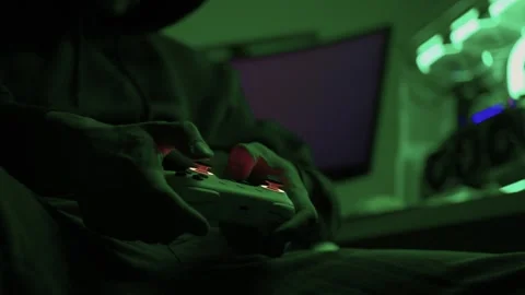 Gamer gripping red controller in dark hoodie, lowlit room with neon green PC and Stock Footage 347874817