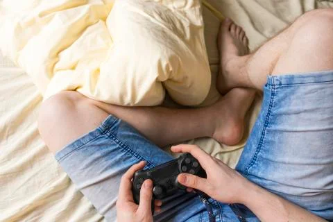 Gamer man sits in rumpled bed with wireless gamepad in his hands Stock Photos