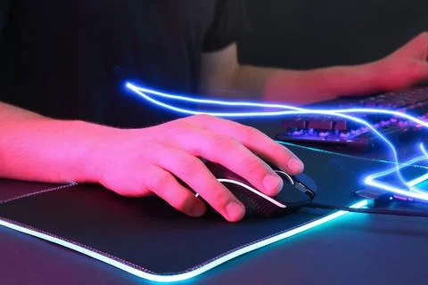 Gamer using modern wired computer mouse in neon lights at dark table, closeup Foto stock