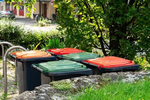 Garbage cans with multi-colored plastic lids stand in an area with green spaces. Stock Photos