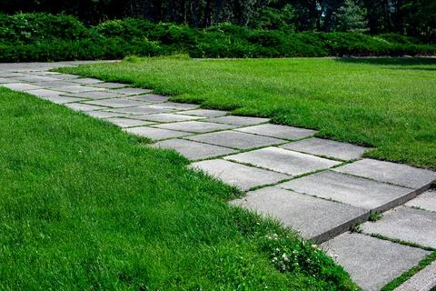 Garden path made of square tiles overgrown with grass in a park. Stock Photos