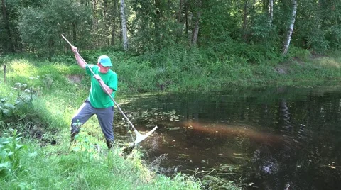 Gardener man with rake tool cleans pond water from grass and sludge. 4K Stock Footage 59338603