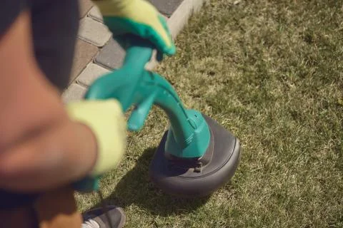 Gardener in sneakers and colorful gloves is mowing green grass with handheld Stock Photos