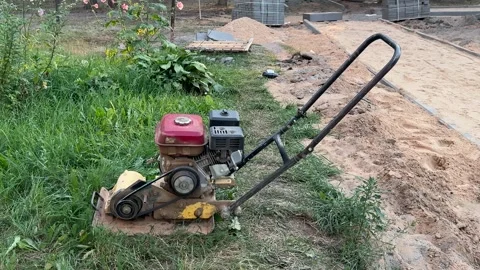 A gasoline plate compactor stands on the grass next to sand  Video stock 341951886