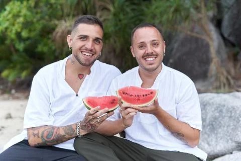 Gay couple looking at camera while holding a piece of watermelon on a beach Stock Photos