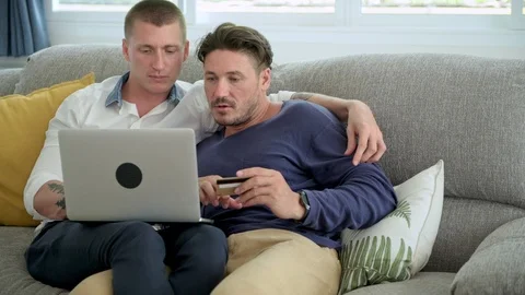 Gay couple relaxing on couch using laptop computer. Keying in credit card Stock Footage 105314878
