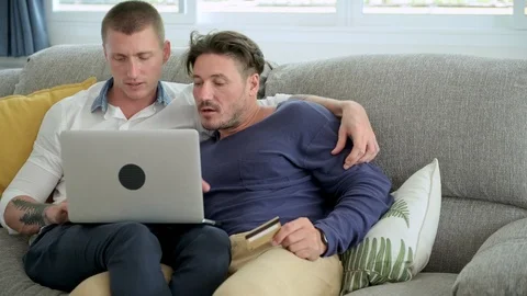 Gay couple relaxing on couch using laptop computer. Complete purchase and kiss. Stockbeeldmateriaal 105315692
