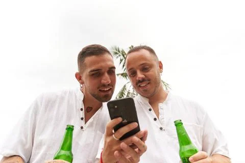Gay couple using the mobile while holding a bottle of beer outdoors Stock Photos