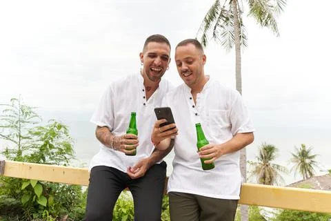 Gay couple using the mobile while drinking beer in a terrace overlooking the sea Stock Photos