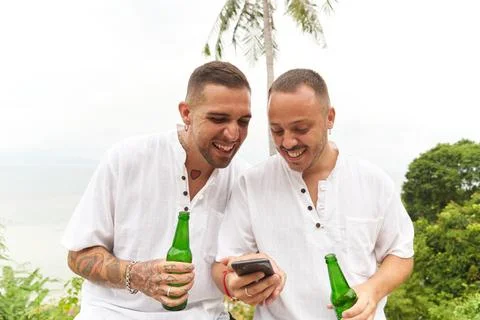 Gay couple in white using a mobile while holding beer next to a tropical beach Stock Photos