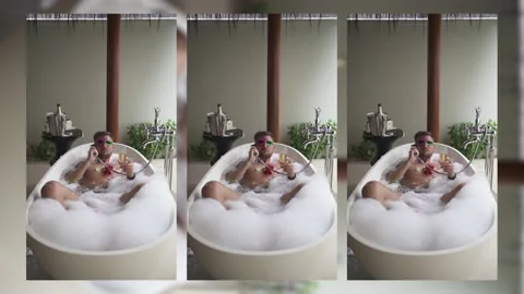 A gay guy is lying in a bubble bath with flower petals holding a glass Stock Footage 150934455