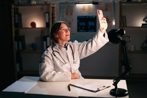 General practitioner doctor making selfie using smartphone working late at ni Фото