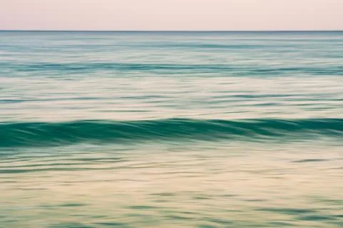 A gentle ocean wave flows under soft pastel-colored skies during calm light. Stock Photos