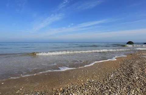 Gentle waves lap a pebble beach under a vast blue sky with wispy clouds 写真素材
