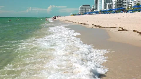Gentle waves reaching sandy Miami beach with sunbathers and blue beach umbrellas Stock-Footage 290455525