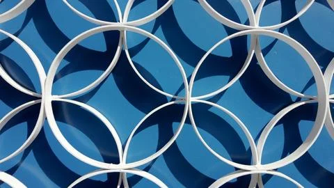 Geometric white metal circle pattern casting sharp shadows on a vibrant blu.. Stock Photos