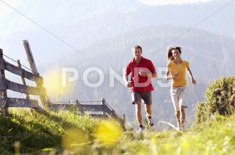 Germany, bavaria, young couple jogging by wooden fence, mountains in backgrounds Stock Photos