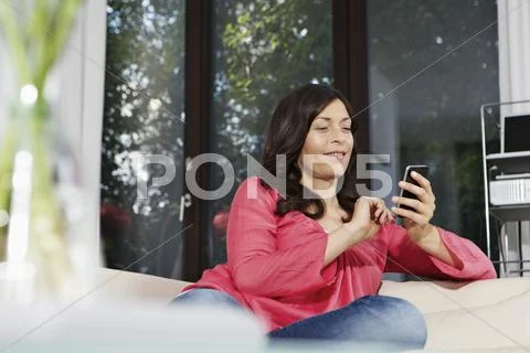 Germany, Berlin, Mature Woman Using Mobile On Sofa, Smiling