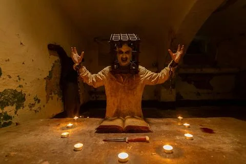 Ghost with cage on his head reading spells next to crucified body in basement Stock Photos