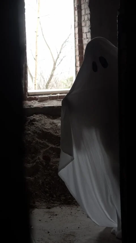 Ghost in white sheet peeks out from behind wall near window, playing hide and Stock Footage 320248182