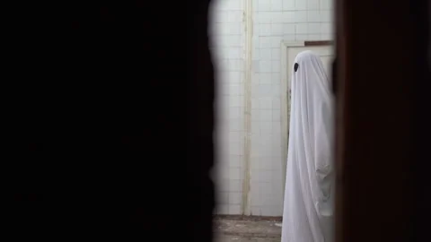 Ghost in a white sheet walks in an abandoned house looks at the camera through Video stock 244455528