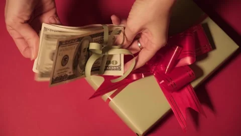 Gift box and stack of dollars on red background, representing cash gifts for Stock Footage 322543091
