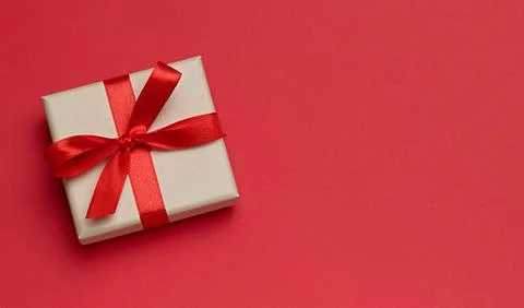 Gift box in beige packaging with red satin bow on a red background, top view Stock Photos