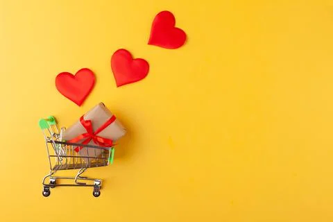 Gift box with red ribbon inside mini grocery cart, red hearts on yellow Stock Photos