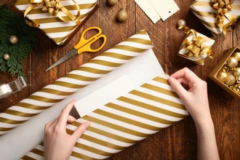 Gift wrapping. Hands wrap a box in striped gold and white paper on a brown .. Stock Photos