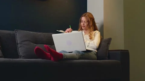 Ginger student with freckles is using a laptop doing her homework on a couch at Stock Footage 155372993