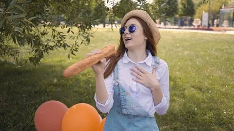 Ginger young girl singing using a baguette as a microphone in park Stockbeeldmateriaal 95247120