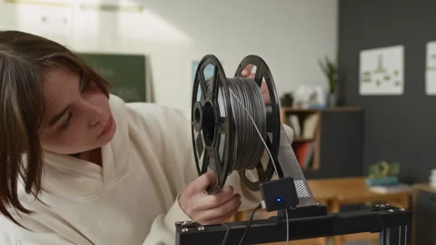 Girl Adjusting Gray Spool of Plastic on Holder of 3D Printer in Classroom Stockbeeldmateriaal 335689306