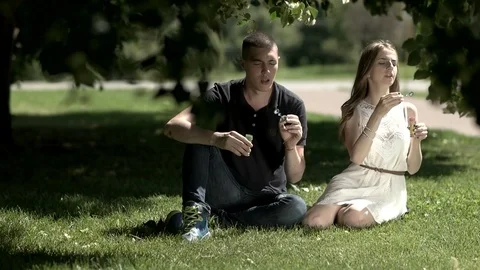 Girl and boy blow the soap bubbles while sitting on the grass Stock Footage
