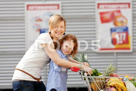 Girl and mother pushing shopping cart together outdoors Stock Photos