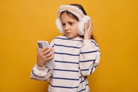 A girl appears surprised while using her smartphone, wearing fluffy earmuff.. Stock Photos