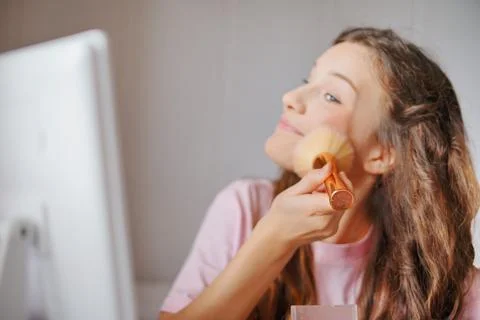 Girl applying makeup while looking at a computer screen in a cozy room with.. Fotos de archivo