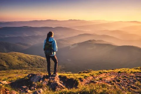 Girl with backpack on mountain peak with green grass at sunset Stock Photos