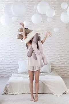 A girl in beige pajamas throws up a stack of silk beauty pillows in the bedroom Stock Photos