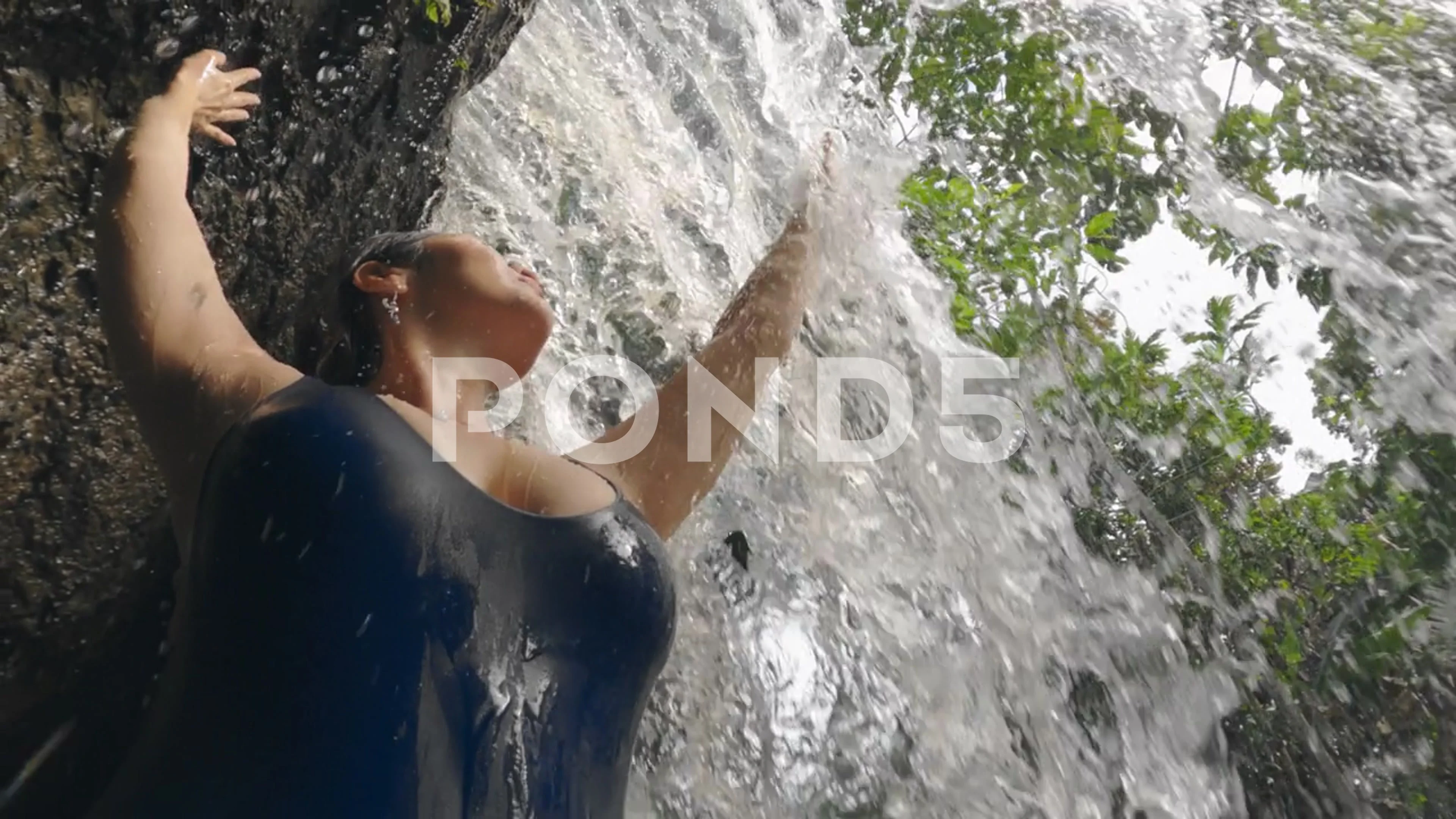 A girl with big breasts in blue swimsuit touching powerful waterfall stream  on